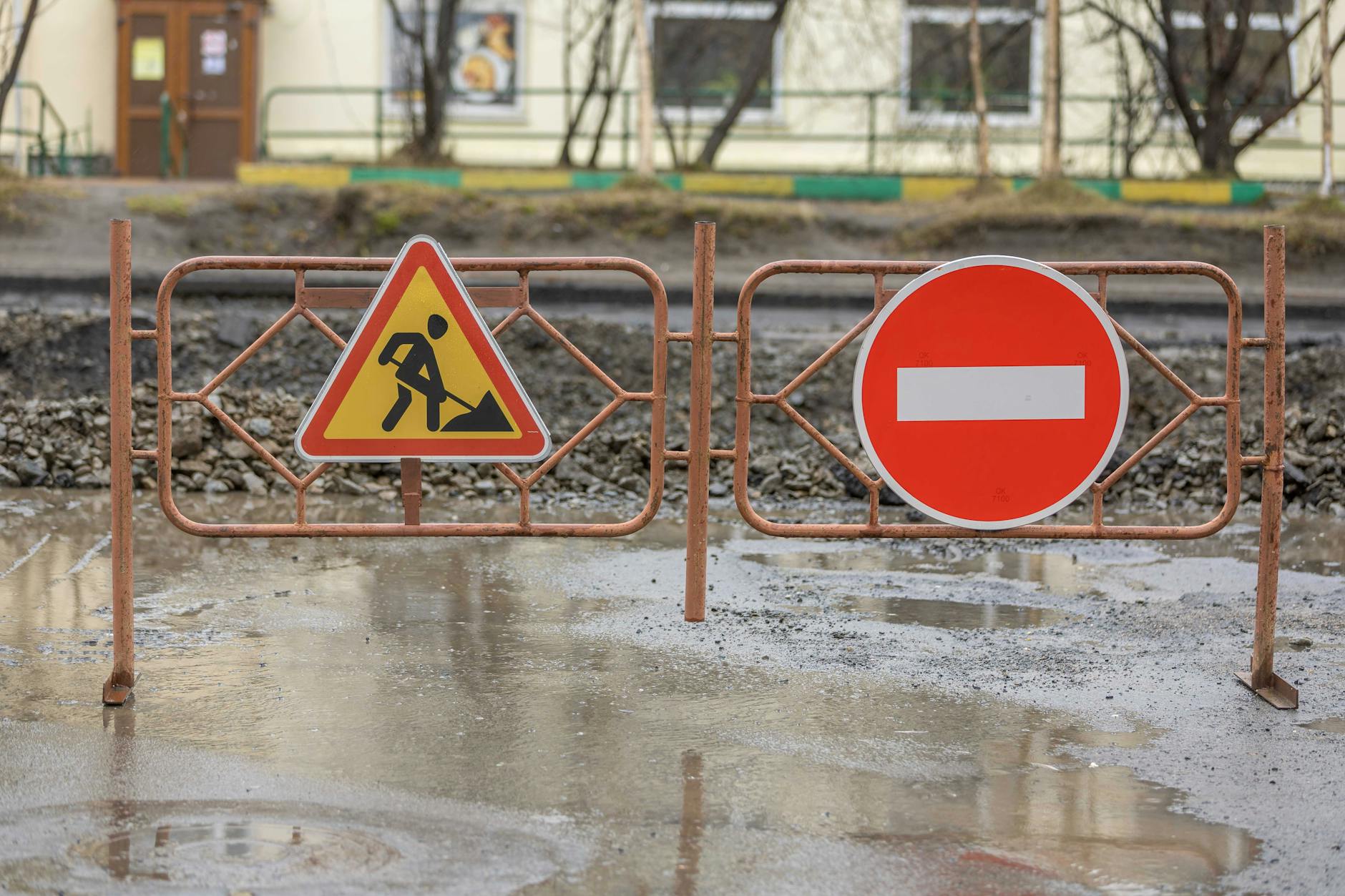 Directional wayfinding signs on a construction site fence pointing to site office and emergency muster point, corflute panels on metal posts