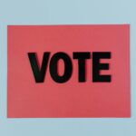 Landscape photo of a row of corflute election campaign signs on a wire fence at a polling place, bright outdoor setting, no identifiable can