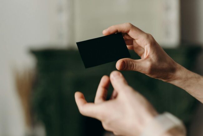 A close-up of a matte black business card with a holographic foil logo catching the light, showing the rainbow colour shift against the dark