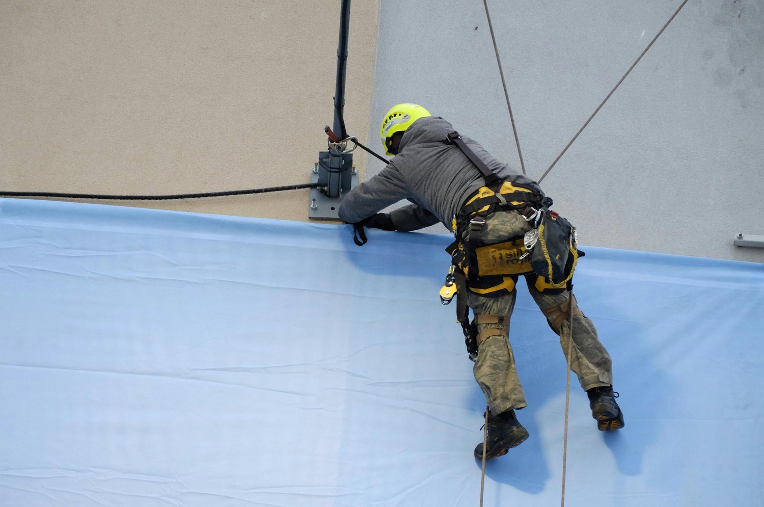 Banner installation specialist on a lift line tensioning a large blue stretch fabric banner against a building, illustrating the professional install techniques discussed below.