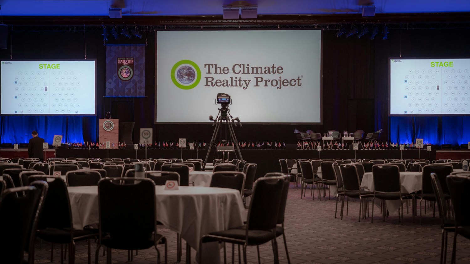 Corporate event hall with branded media walls displaying The Climate Reality Project logo across a wide central panel, suitable for sponsor grid layouts.