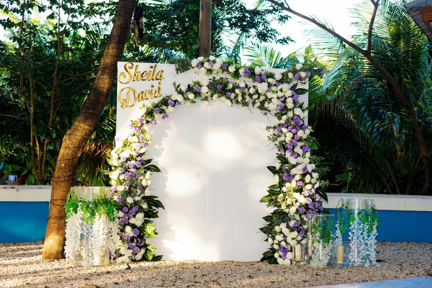 White rectangular printed wedding media wall with Sheila and David in gold script and a lush floral border, set in an outdoor garden ceremony.