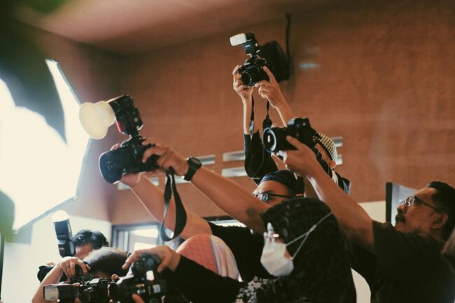 Press photographer scrum at a high-profile media event, cameras raised at presenter height — the working distance media wall sizing accounts for.