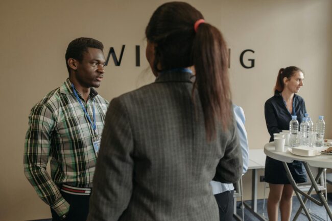 two professionals at an industry networking event exchanging business cards across a table with name badges visible in the background
