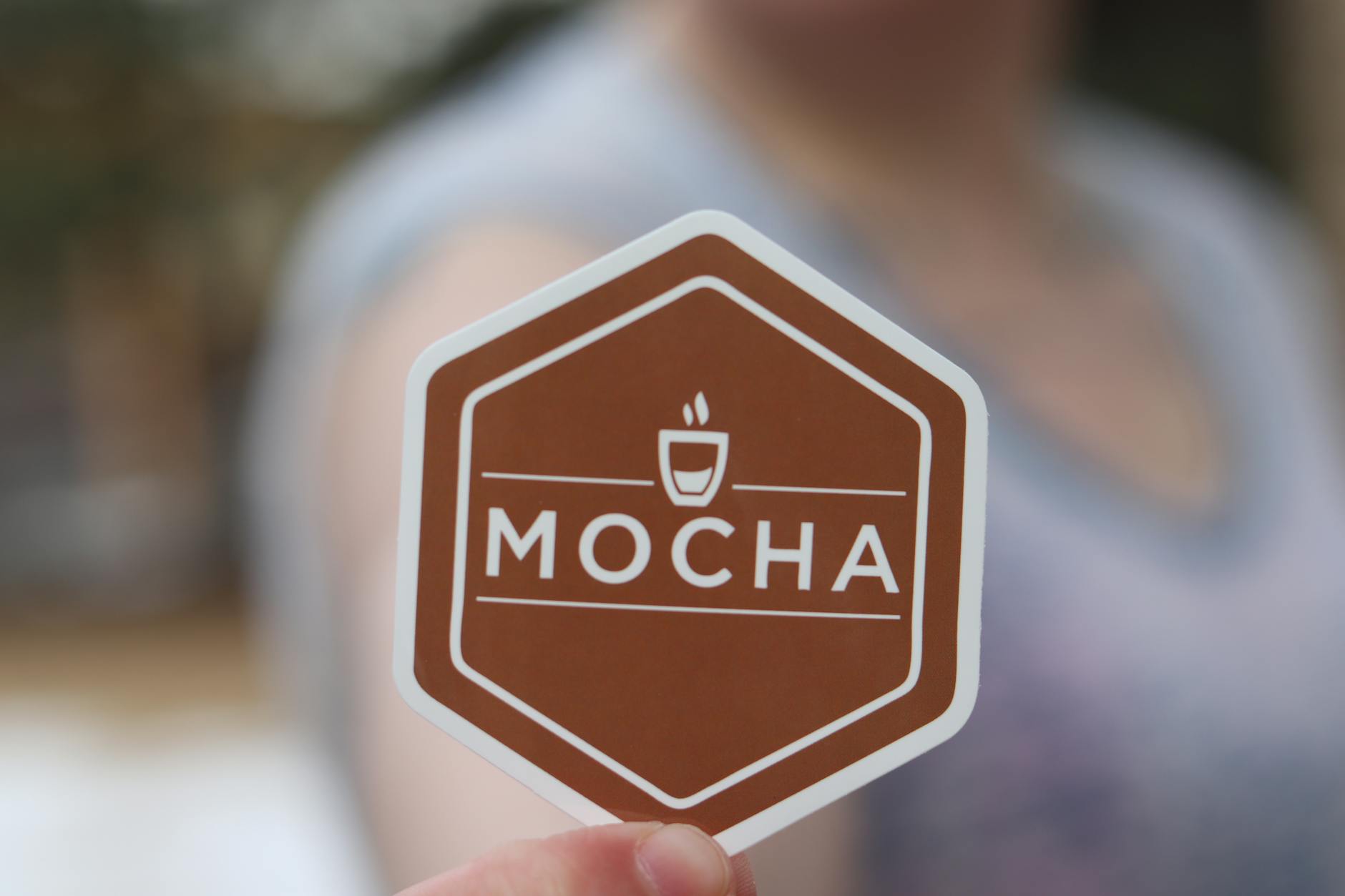 Close-up of a branded die-cut logo sticker being peeled from its individual white backing at a trade show table, with branded merchandise vi