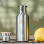 a brushed stainless steel reusable water bottle with a bamboo cap beside a fresh lemon, dishwasher-safe drinkware styling