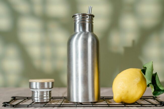 a brushed stainless steel reusable water bottle with a bamboo cap beside a fresh lemon, dishwasher-safe drinkware styling