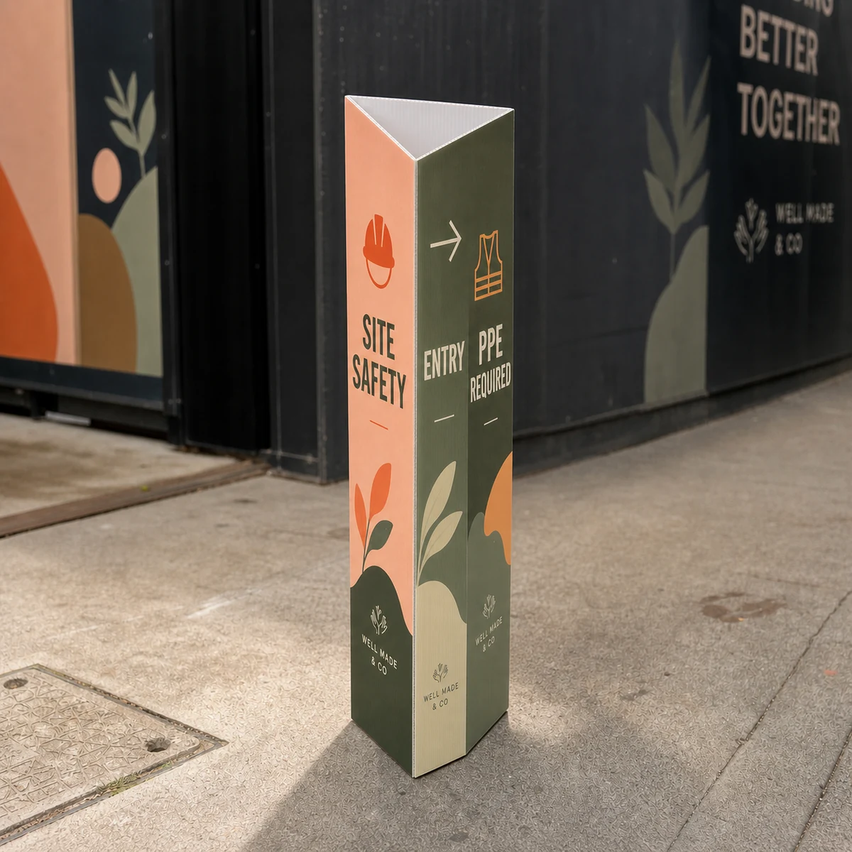 A triangular bollard sign on the ground at a construction site entrance carrying bold site-safety and directional text, a building hoarding softly out of focus behind