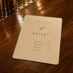 A laminated bar drinks menu on a polished timber bar top with a condensation ring from a nearby glass, the print staying crisp and undamaged, moody low lighting