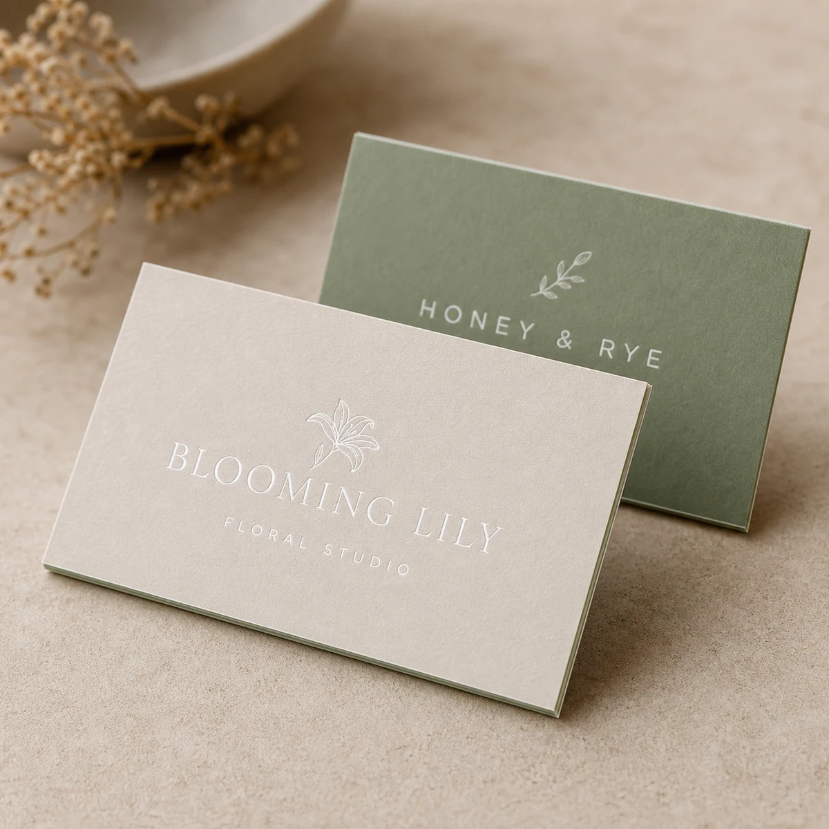 Two thin duplex business cards propped so the slim two-tone two-ply cut edge is visible, a sage-green reverse and a white-ink wordmark on the cream face