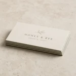 Overhead flat lay of a low stack of thin duplex business cards on light stone with the slim two-tone two-ply bonded cut edge visible, a cream face over a sage-green ply
