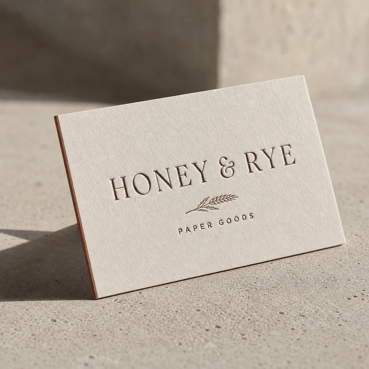 A single thin letterpress duplex business card propped at an angle on concrete in raking light, the debossed impression casting a clear shadow, the fine two-tone two-ply edge visible