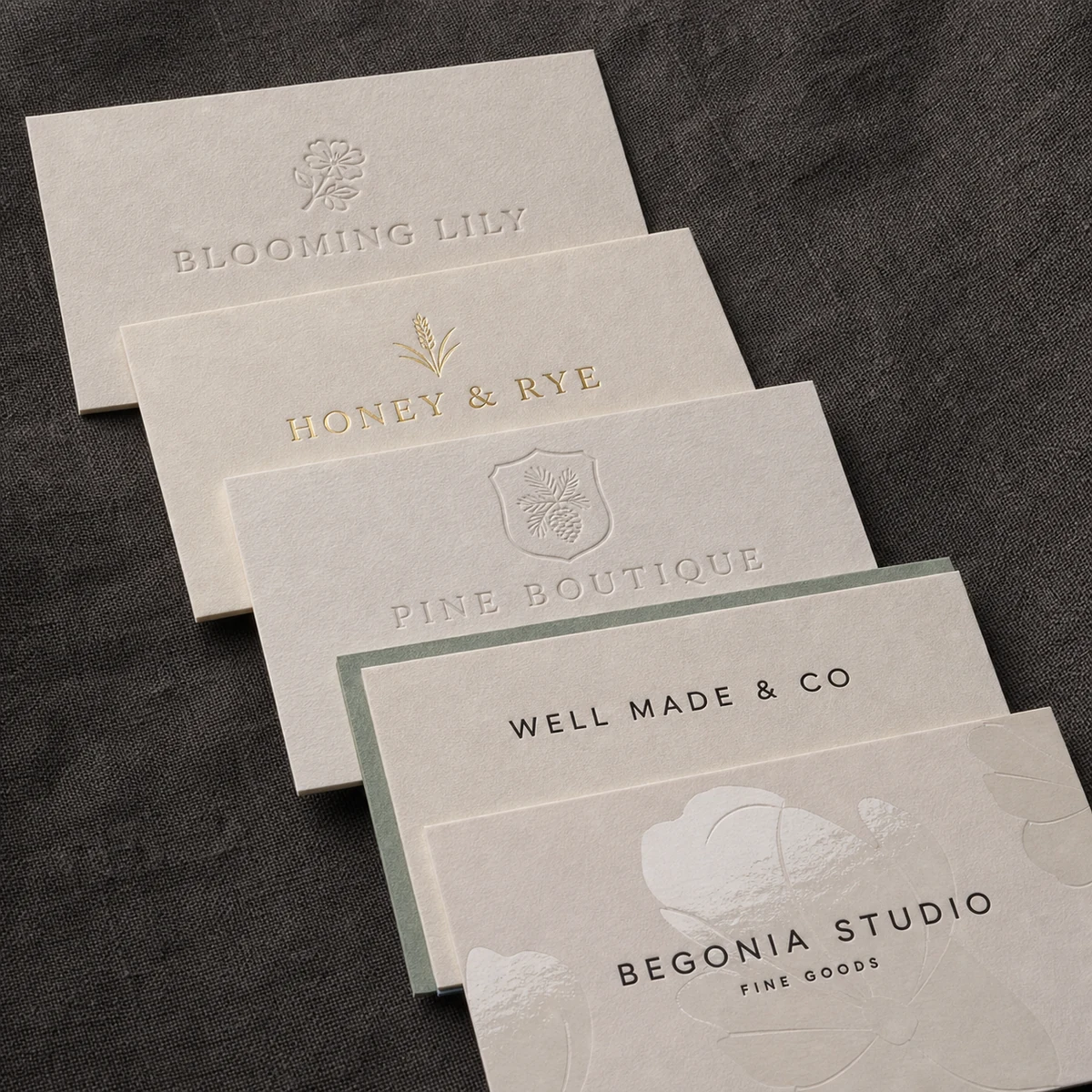 Five premium business cards fanned on dark linen, each a distinct finish: a cotton letterpress blind deboss, a gold foil card, an embossed crest, a thin duplex card and a spot-gloss card