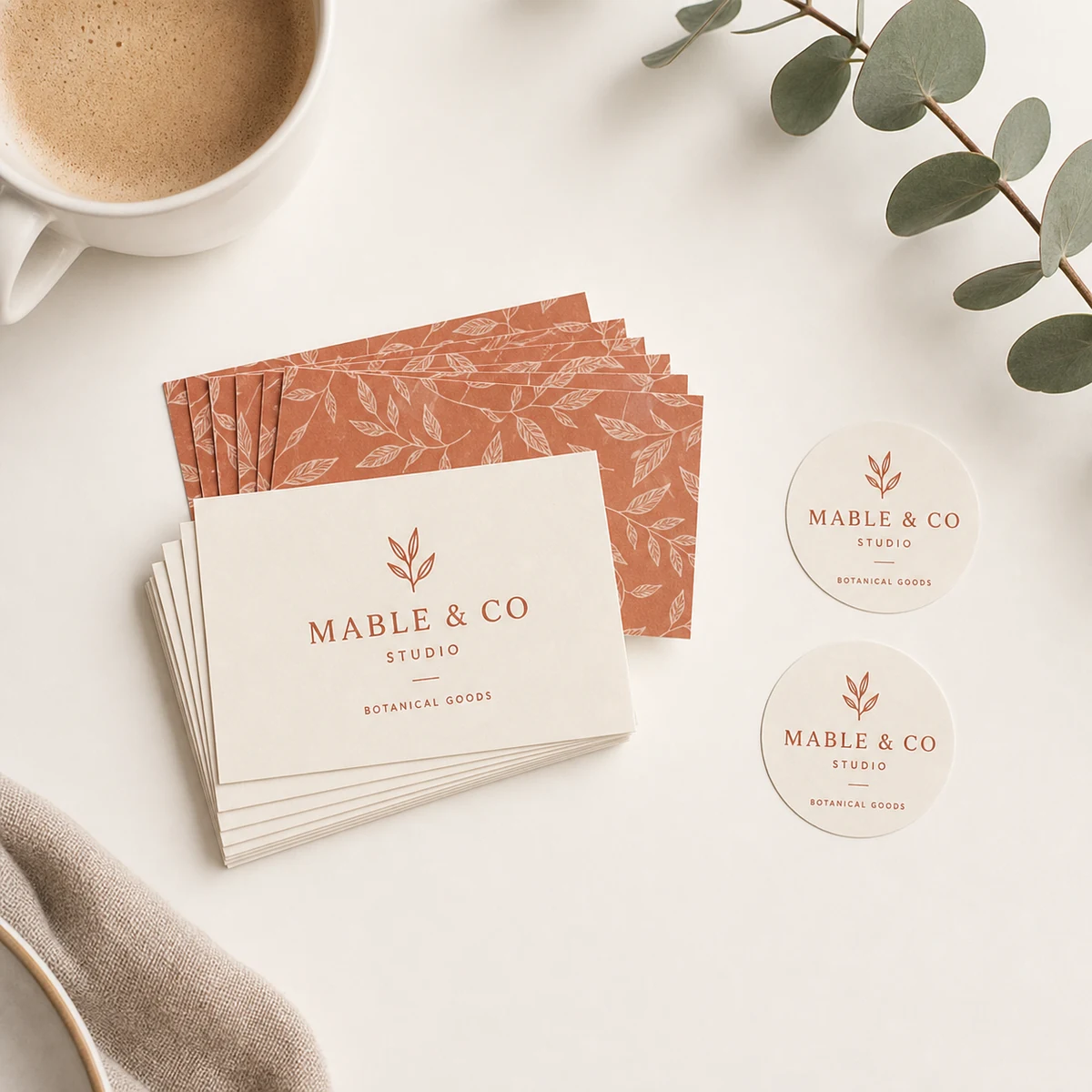Overhead flat-lay of a new-business brand launch set on a clean white desk, a fan of landscape business cards beside two round circle stickers in a warm terracotta and cream MABLE AND CO identity, with a flat-white coffee cup and a sprig of eucalyptus