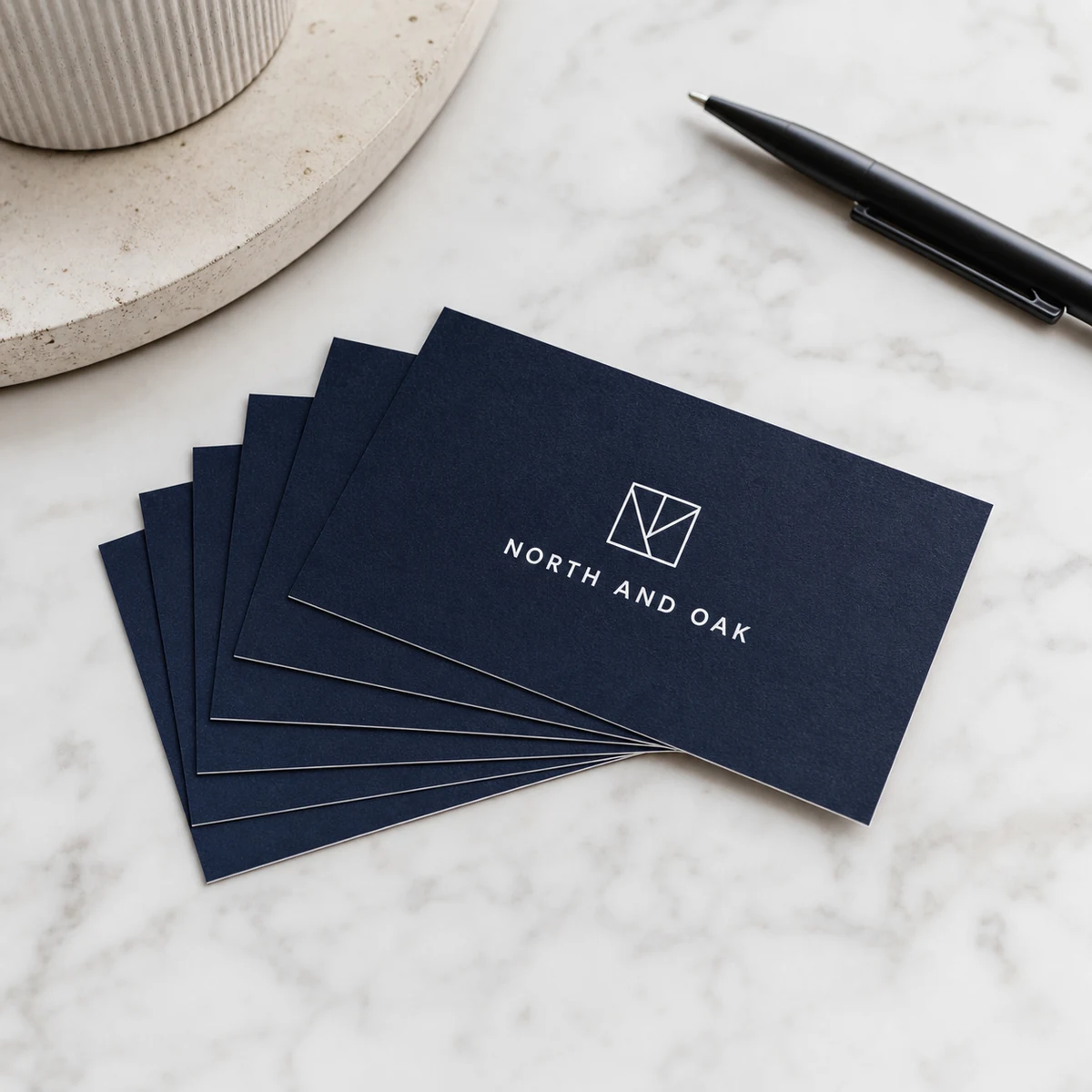Premium standard business cards printed in rich deep navy with crisp white text and a small geometric logo, fanned flat on a white marble surface, showing the clean sharp quality of CMYK digital printing on premium card stock