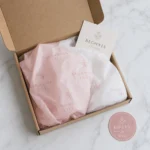 Overhead flat lay of branded blush and white tissue paper with a repeating logo pattern folded inside an open kraft mailer box beside a round sticker seal on marble