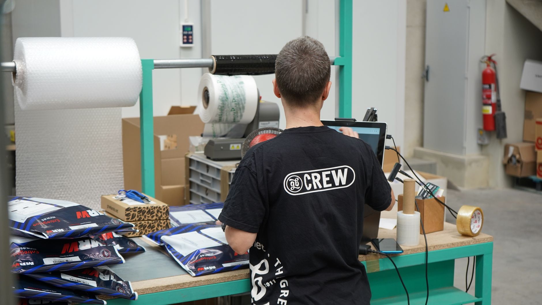 a packing bench with a roll label dispenser, a stack of white cardboard boxes and a peeled branded address label mid-application