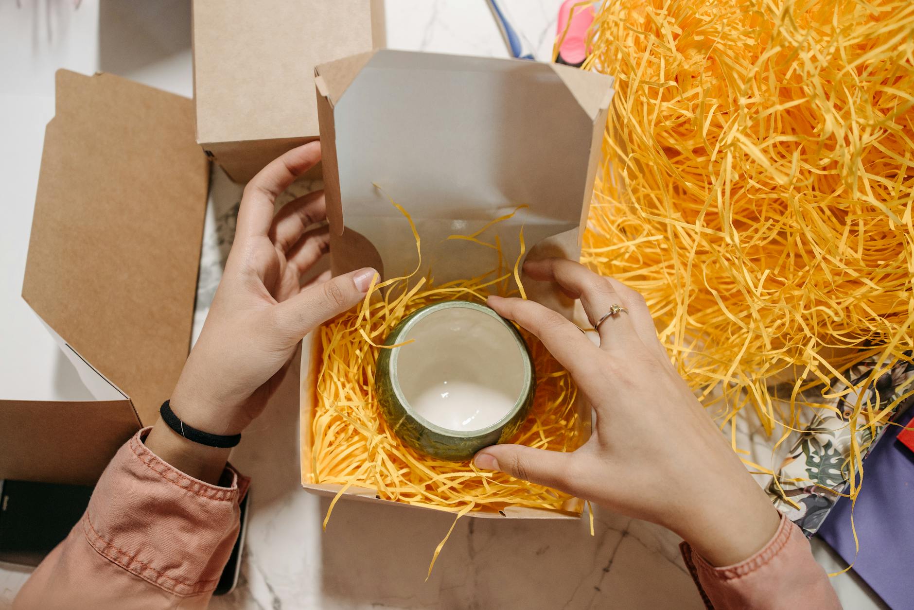 hands packing a ceramic cup into a kraft gift box with shredded paper filler for an online order