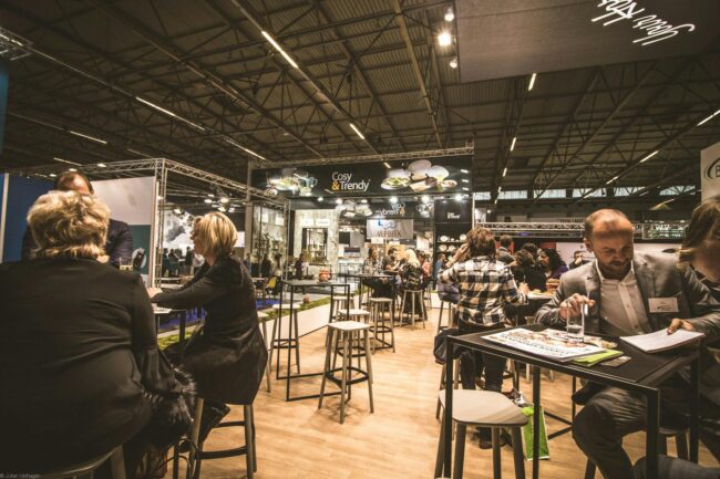 a fully dressed trade show booth with a stretch-fabric media wall backdrop, two pull-up banners on each flank, and a table covered with