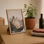 A4 natural oak framed print of a protea standing on its fold-out stand on a timber dining table beside a potted plant, olive oil and folded linen napkins
