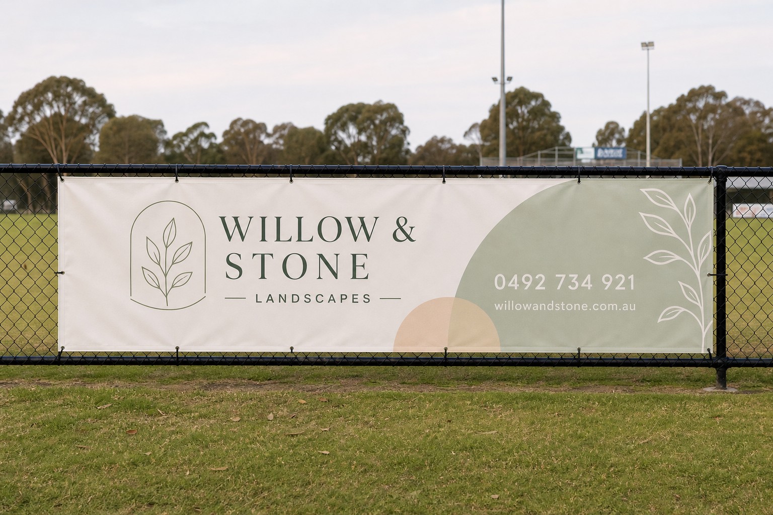 A 3000mm x 1000mm vinyl sponsor banner zip-tied to a chain-link boundary fence at a community oval