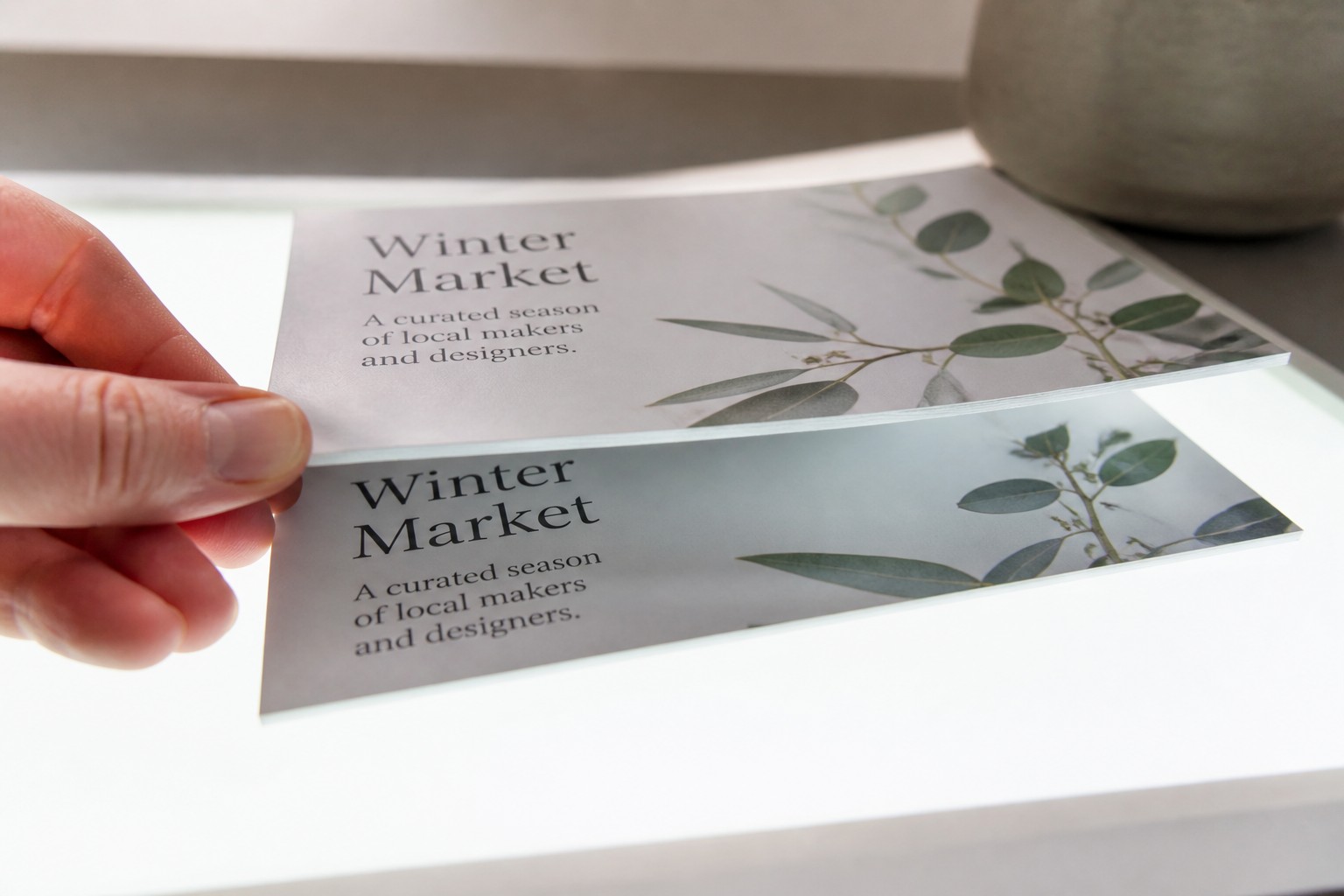 A hand lifts one flyer from a stack on a lightbox, the eucalyptus branch design glowing through the backlit sheets.
