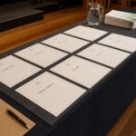 A trestle table with a charcoal runner holds three rows of portrait certificates, folded programs, a water jug, a kraft envelope and pen in a hall.
