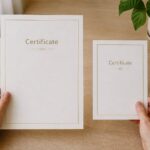 Two hands hold a larger and a smaller portrait certificate side by side, each cream with a fine gold keyline and a short matching gold title.
