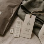 A woven size label, printed care strip and branded hang tag tied with twine rest against folded knitwear, beside a kraft bag and label roll.