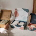 Three custom mailer boxes of stepped sizes on white surface, leftmost kraft with black text mark, center and right featuring full-color patterned designs, all open to show corrugated edges and interior.