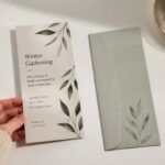 A botanical flyer rests beside its matching envelope on a sunlit desk, held lightly at the corner by hand.
