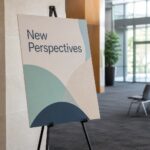 A large printed foam board panel stands on a slim black floor easel in a bright conference centre foyer, its pale edge visible in soft daylight