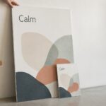 Large and small printed foam boards lean against a pale wall showing sand, terracotta, sage and deep blue layered curves, hand on the larger board.
