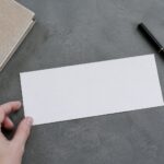 Single DL gift voucher lying face down on a slate grey desk, blank back panel filling the frame, beside black pen and closed notebook.