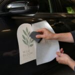 A squeegee smooths a leafy decal onto a dark car door as the backing paper peels away in a shaded garage.