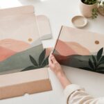 Flat unfolded kraft mailer box blank and fully assembled mailer box with geometric abstract artwork in peach, grey and green tones, both displayed on white work surface with hands holding the assembled box, soft daylight.