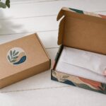 Overhead flat-lay of a kraft mailer box with botanical sticker beside an open full-colour printed mailer box displaying vibrant teal and coral artwork, tissue paper inside, eucalyptus branch styling.