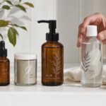 A hand reaches for a clear glass tonic bottle with a leaf print label, beside amber and glass skincare bottles on a bench.