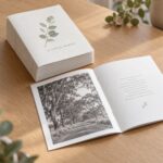 Neat stack of ten A5 portrait order of service booklets on pale oak, off white covers, one open beside with road photo