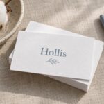 A stack of white cotton business cards on oatmeal linen with raw cotton bolls, a speckled dish, and a grey pen.