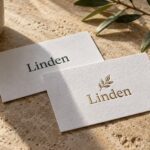 Two white textured cotton business cards overlap on travertine stone, one pressed green name, one raised gold foil name and leaf motif.