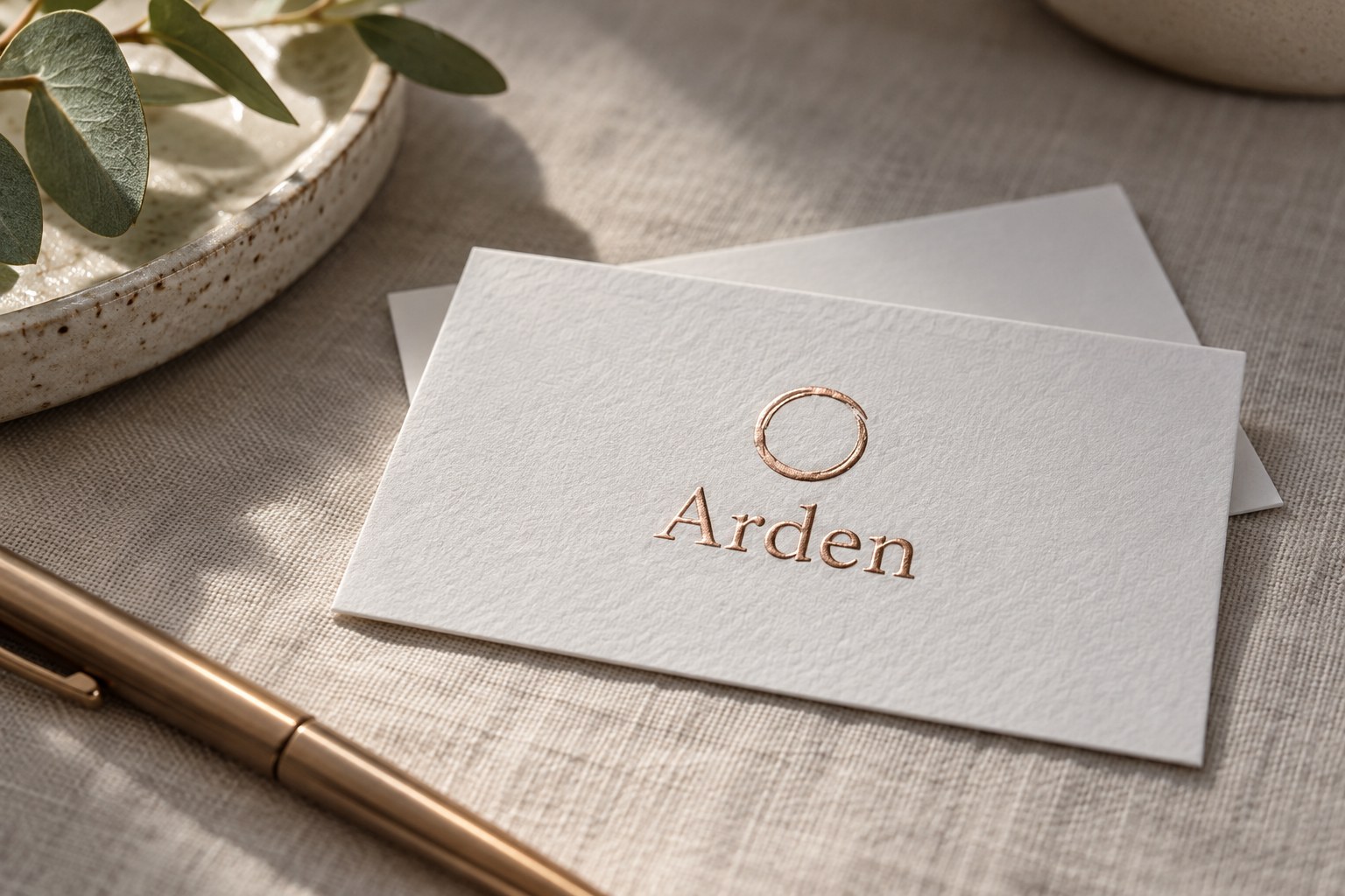 One white textured cotton business card on pale grey linen, its raised rose gold circle and name casting soft side shadows, with a second card behind.