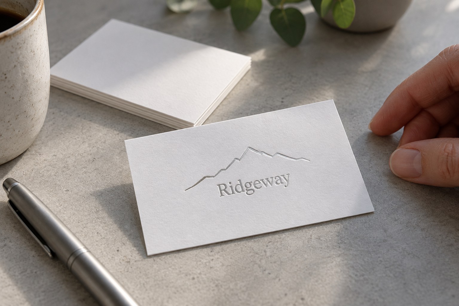 One white cotton business card on pale grey concrete with a silver foil mountain ridge line drawing and name pressed into the card.