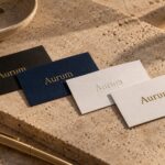 Four business cards fanned on a warm travertine plinth in low sunlight, showing gold finishes on matte black, navy textured, and white cotton stock.