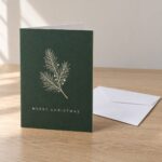 Folded A6 corporate Christmas card slightly open on pale oak desk, cream fir sprig front, plain white envelope behind