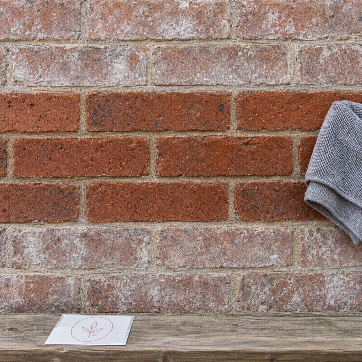 Red brown brick wall half wiped clean, pale chalky film above, grey microfibre cloth and unapplied sticker on timber ledge