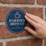 Hand pressing round deep blue high tack sticker with cream serif capitals flat onto red brick wall with grey mortar joints