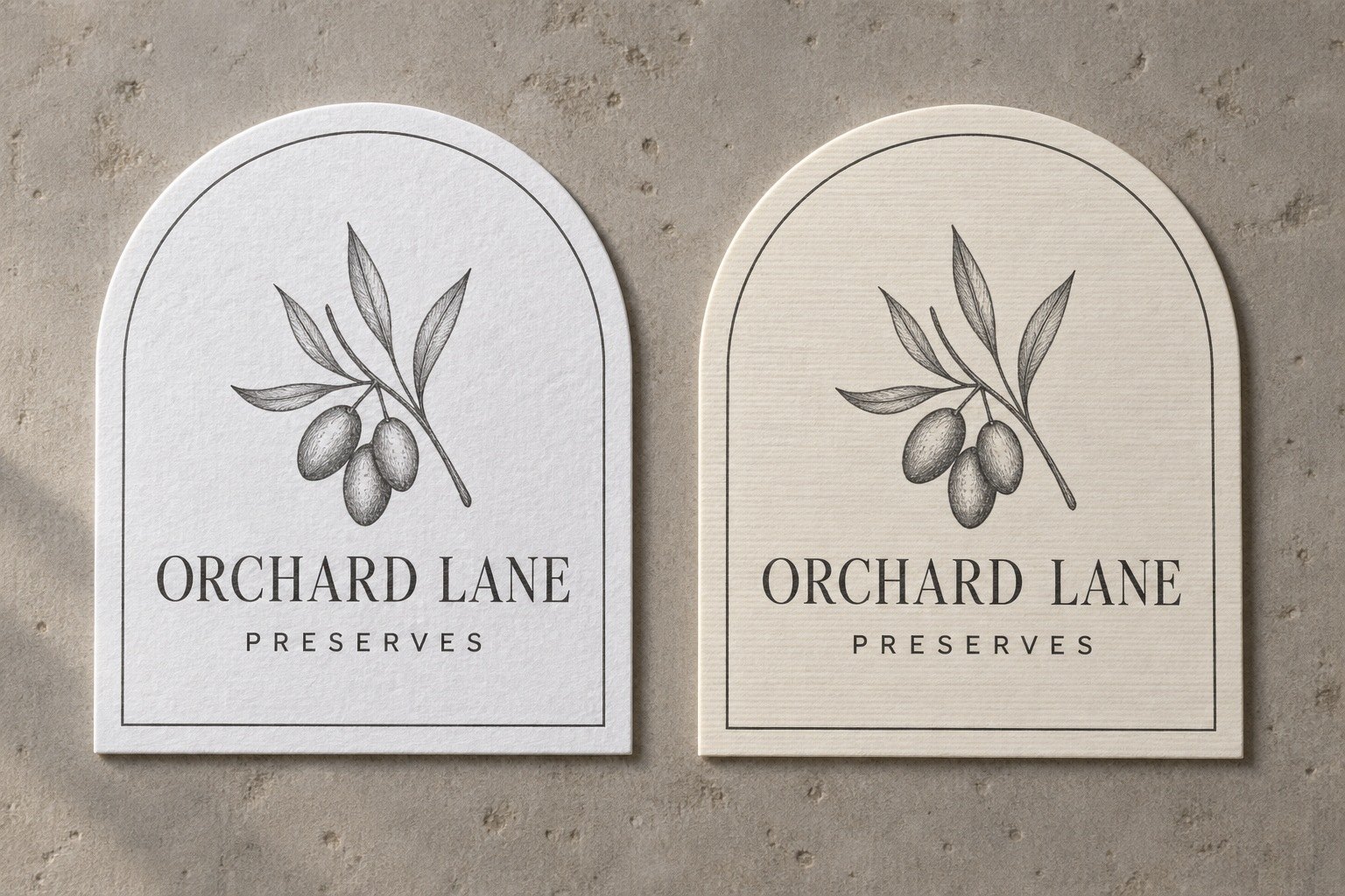 Two arch topped paper stickers with charcoal olive sprig, felt and linen textures side by side on warm grey stone