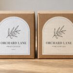 Two identical kraft brown gift boxes with textured stickers, one white felt label and one cream linen label on pale oak table