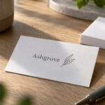 White cotton business card with charcoal pressed lettering and a leaf line drawing on a pale oak desk beside blank white cards and blurred leaves.